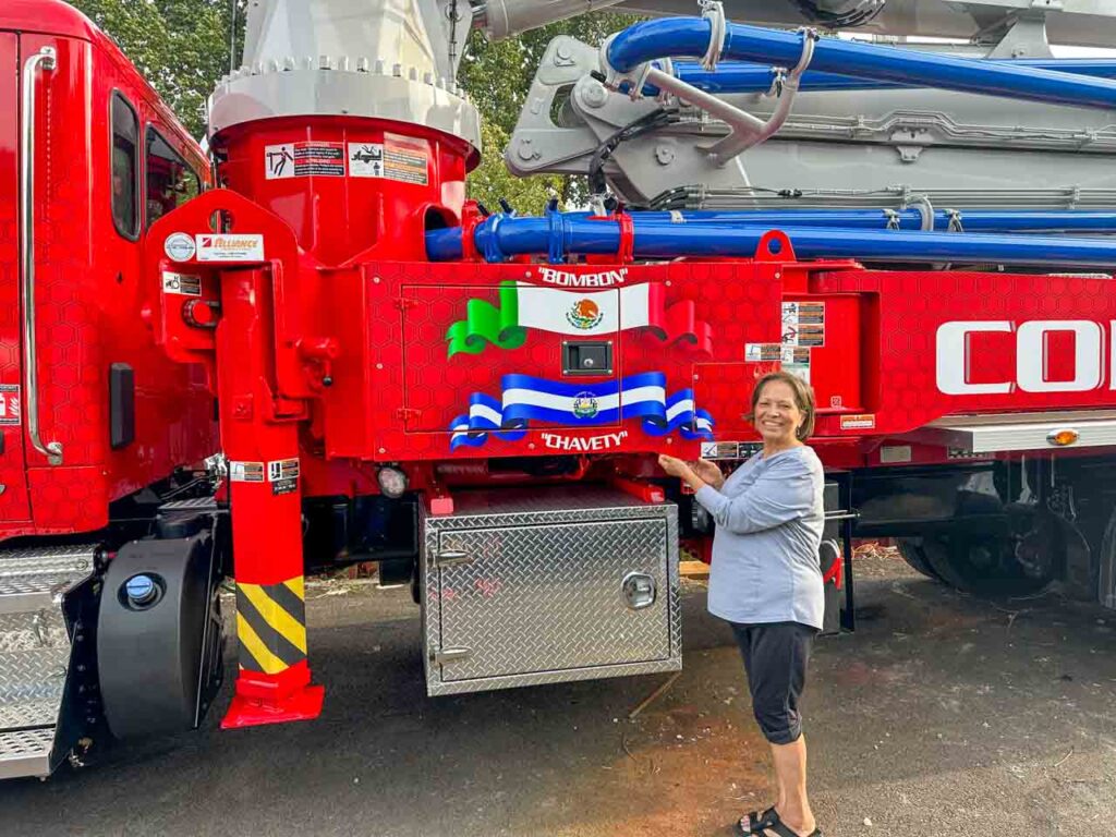 A woman standing next to a red concrete pump truck, smiling and pointing at the vehicle.